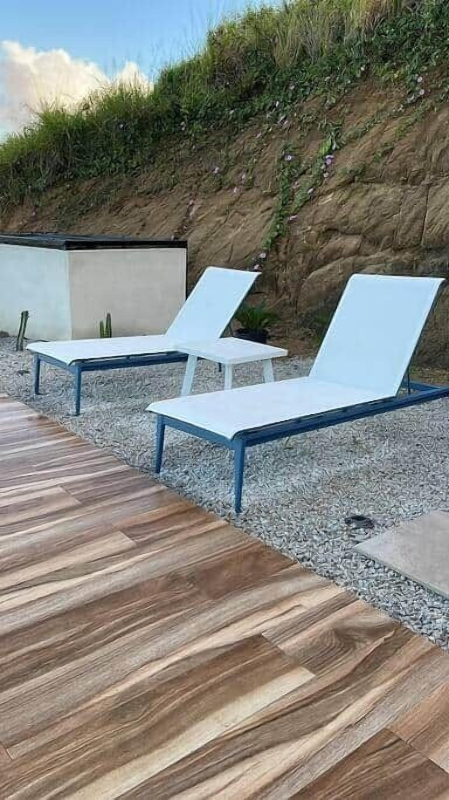 Two white lounge chairs with blue metal frames, positioned on a wooden deck with a rocky backdrop.