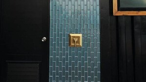 Un bâtiment avec une porte noire, un mur recouvert de carreaux bleus et orné d’une sonnette dorée, ainsi qu’un cadre de fenêtre.