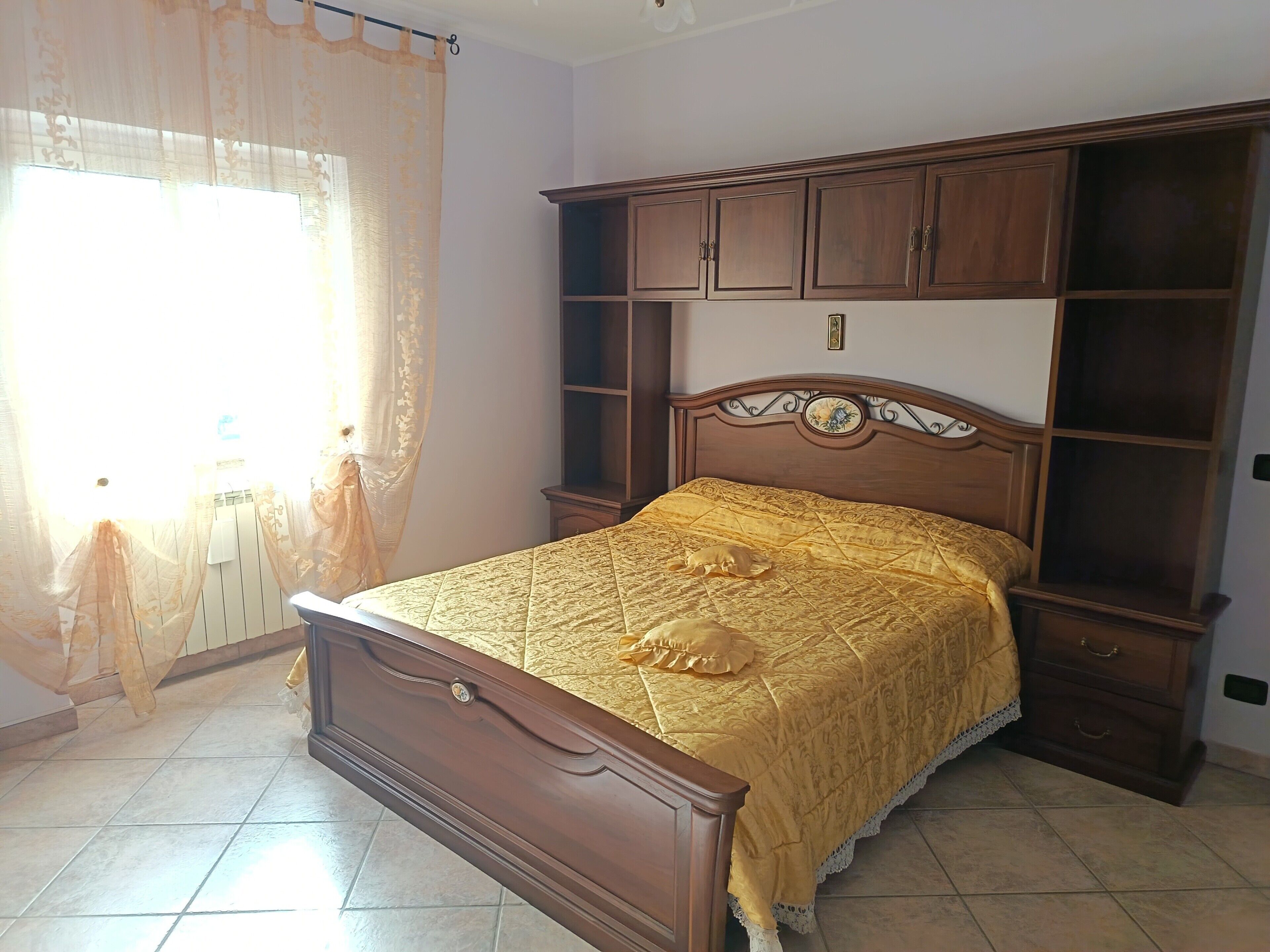 A bedroom with a wooden bed, a nightstand, a wardrobe, and a window with sheer curtains.