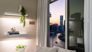 A modern living room with a city view, a white sofa, a plant, and a shelf with books and a sculpture. - Waterfront Escape | Burj & Canal View | Pool+Sauna (Dubai)