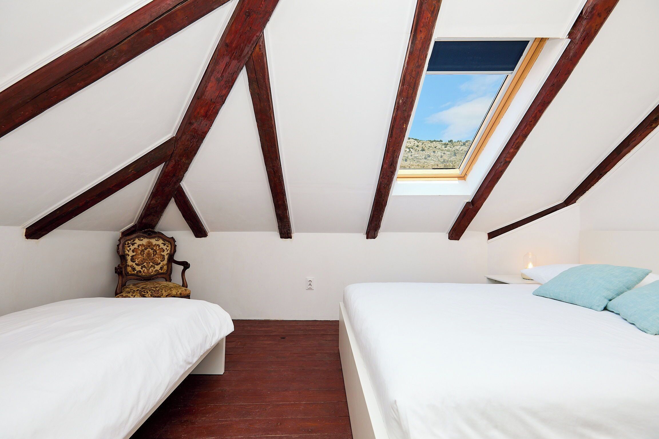 A bedroom with two beds, a skylight, wooden beams, a chair, and a nightstand.