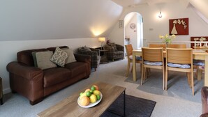 A cozy living room with a brown leather sofa, a wooden coffee table with fruit, a dining area with wooden chairs and a table, and a kitchen in the background.