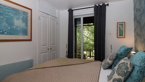 A bedroom with a bed, a painting, a door, and a view of greenery outside.