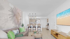 A modern living room with a sofa, coffee table, and a painting of a beach. - Stunning 1BR Apartment with Balcony & Pool (Hallandale Beach)