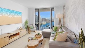A modern living room with a large window, a sofa, a coffee table, and a TV. - Stunning 1BR Apartment with Balcony & Pool (Hallandale Beach)