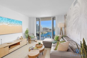 A modern living room with a large window, a sofa, a coffee table, and a TV. - Stunning 1BR Apartment with Balcony & Pool (Hallandale Beach)