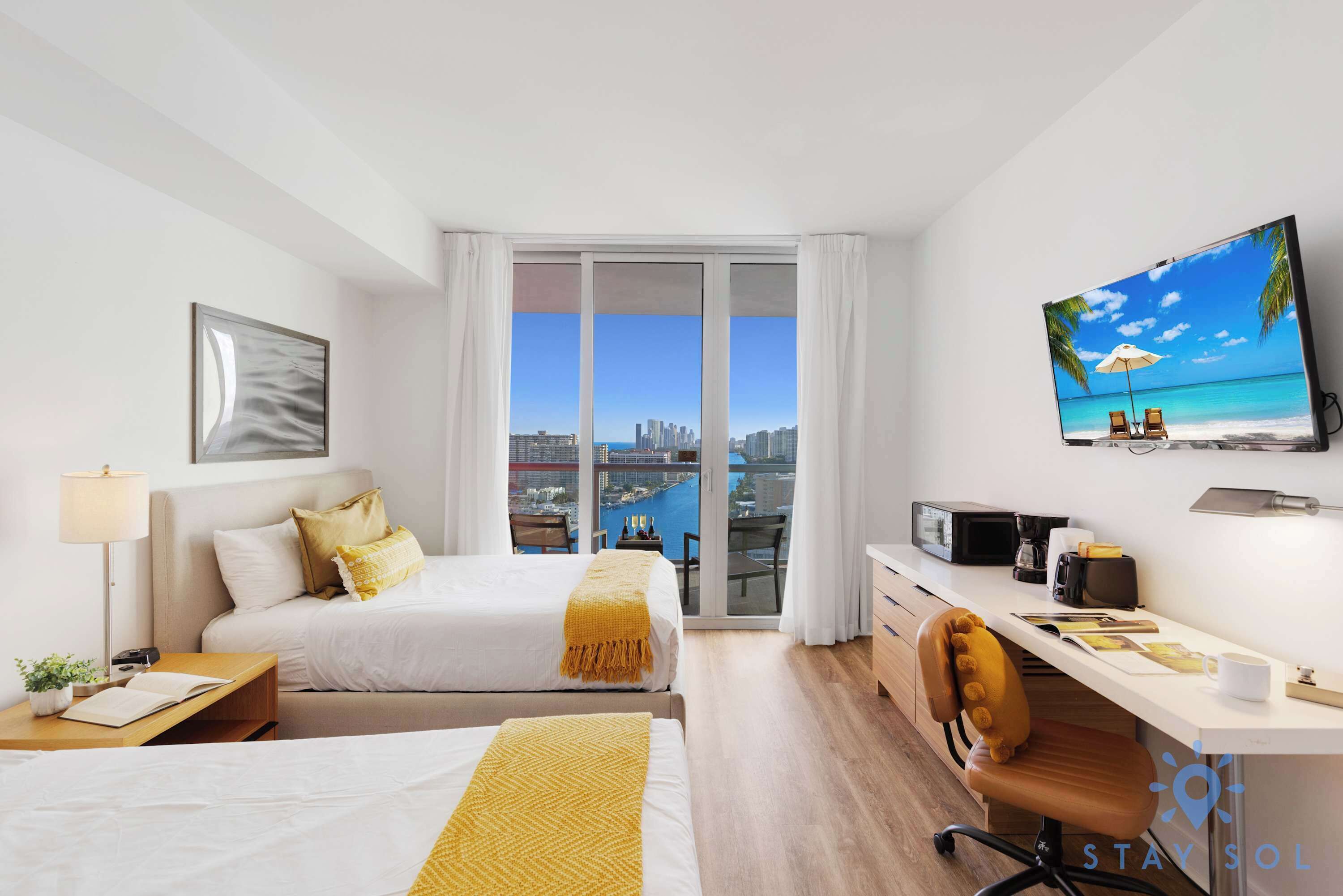 A hotel room with a bed, desk, chair, TV, and a balcony with a city view.