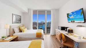 Ein Hotelzimmer mit Bett, Schreibtisch, Stuhl, Fernseher und einem Balkon mit Blick auf die Stadt.