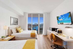 A hotel room with a bed, desk, chair, TV, and a balcony with a city view. - Amazing Studio w/ Pool and Gym (Hallandale Beach)
