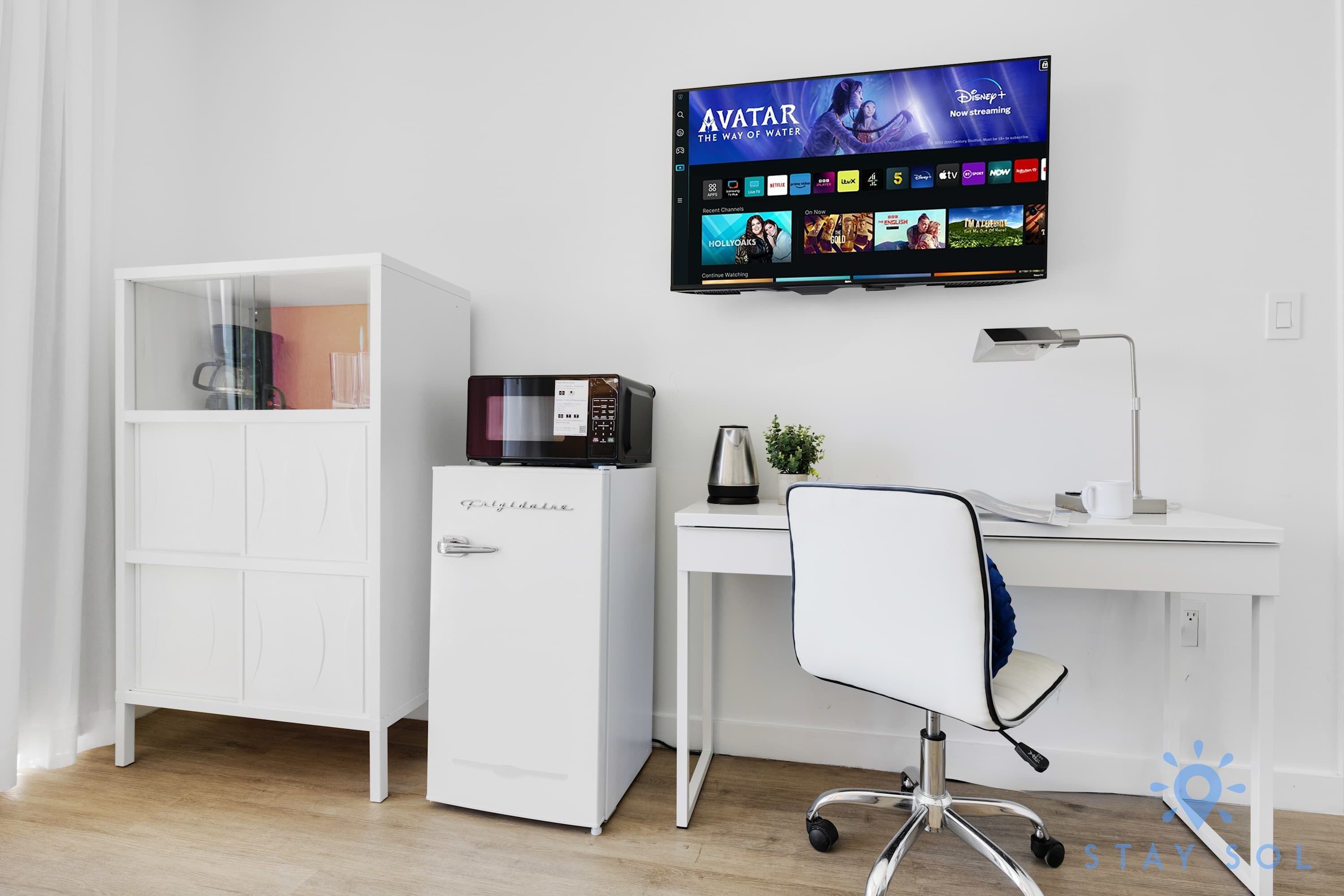 Un espacio de oficina moderno con un escritorio blanco, una silla blanca, un archivador blanco, un microondas y un televisor de pantalla plana mostrando la interfaz de Disney+.