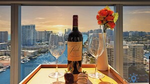 A bottle of wine, two glasses, and a vase with flowers on a tray by a window with a city view. - Beautiful Studio w/ Stunning Views + pool + gym (Hallandale Beach)