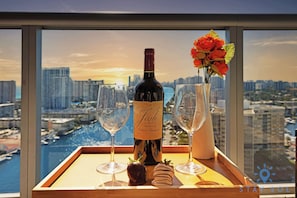 A bottle of wine, two glasses, and a vase with flowers on a tray by a window with a city view. - Beautiful Studio w/ Stunning Views + pool + gym (Hallandale Beach)