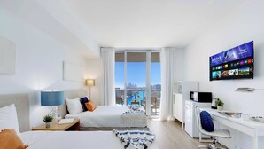 A modern hotel room with two beds, a desk, a TV, and a balcony with a city view. - Beautiful Studio w/ Stunning Views + pool + gym (Hallandale Beach)