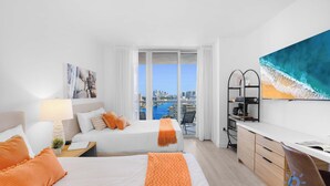 A hotel room with two beds, a desk, a chair, a balcony with a city view, and a painting of a wave. - Steps to Beach Studio, Balcony, Pool & Gym Access! (Hallandale Beach)