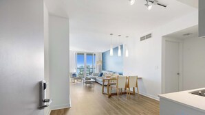 A modern hotel room with a wooden floor, a dining area with a table and chairs, a sofa, and a large window offering a city view. - Bright Apartment | Panoramic View | Resort Vibe (Hallandale Beach)