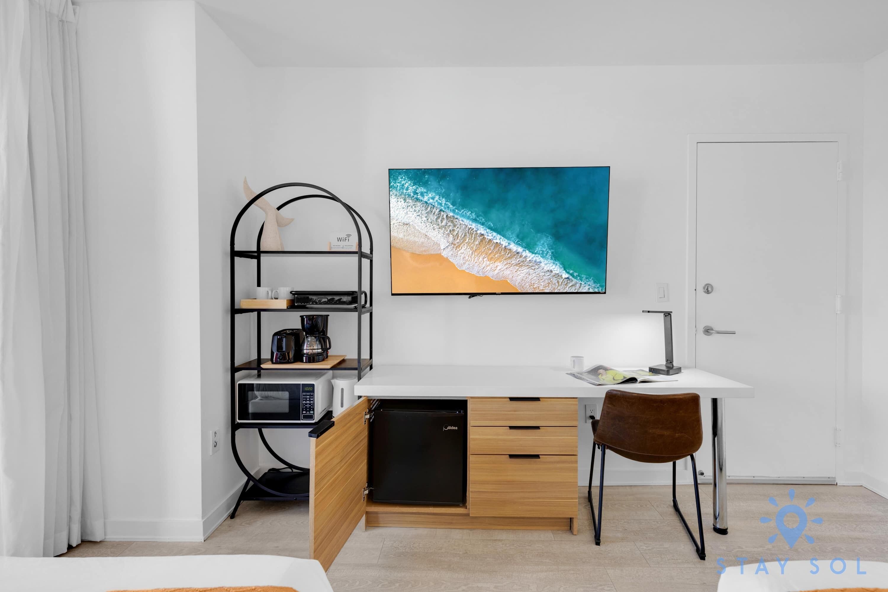 Une chambre d’hôtel compacte comprenant un coin cuisine, un lit, un bureau avec une chaise et un grand tableau sur le thème de la plage accroché au mur.