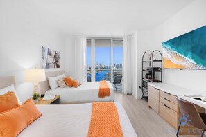 Habitación de hotel con dos camas, un escritorio, una silla, un balcón con vista a la ciudad y un cuadro de una ola.