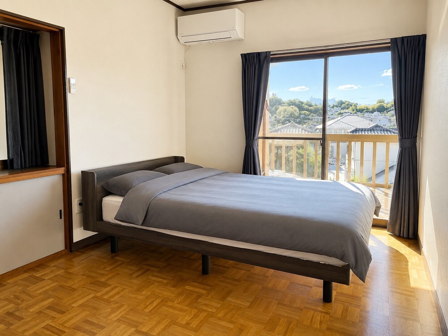 A bedroom with a bed, wooden flooring, a window with curtains, and an air conditioning unit.