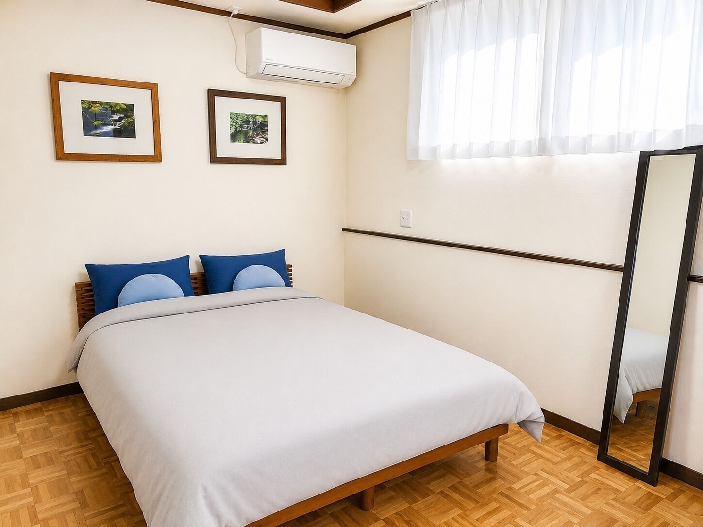 A bedroom with a bed, two framed pictures, an air conditioning unit, a window with curtains, and a mirror.