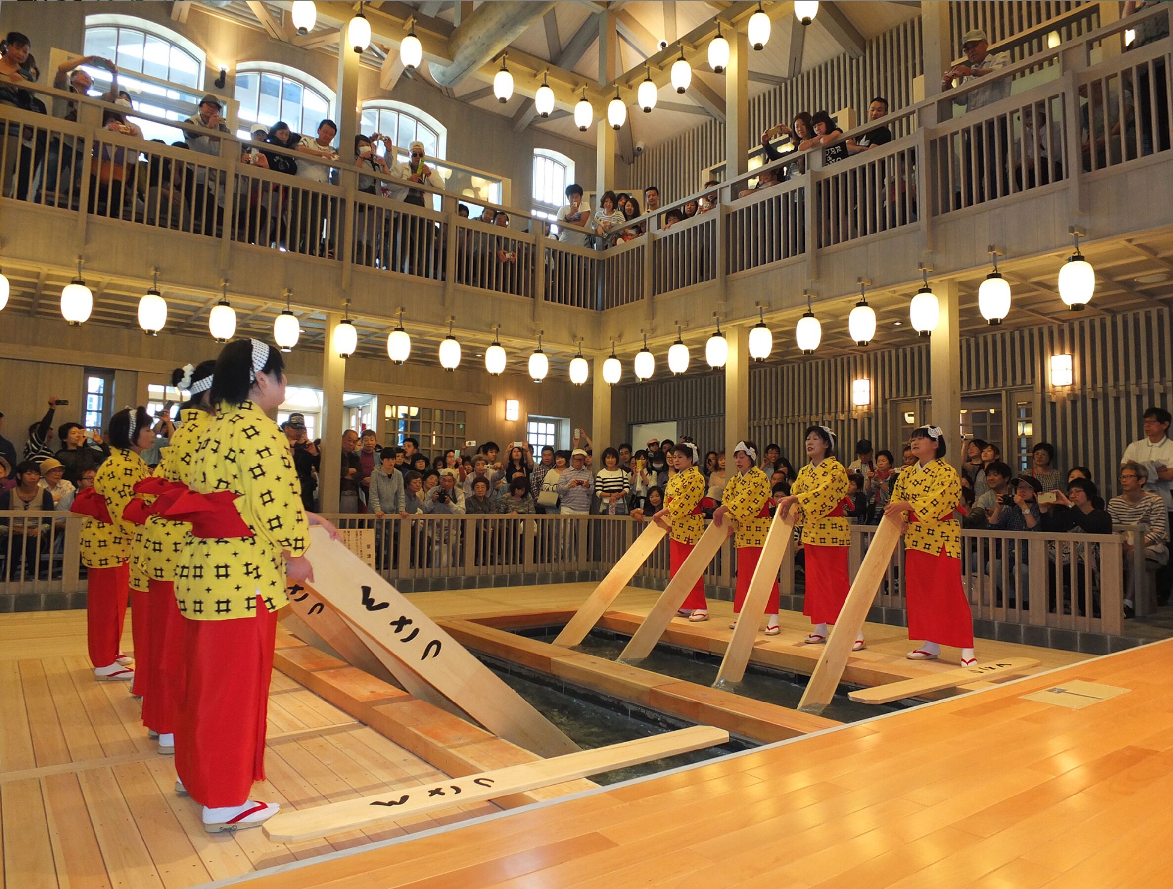A traditional Japanese event with participants in yellow and red attire, a wooden stage, and an audience in a multi-level indoor setting.