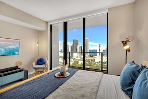 A modern bedroom with a large window, a bed, a chair, a TV, and a bedside table with a bottle and glasses. - Modern Stylish 1BR Apartment (Miami)