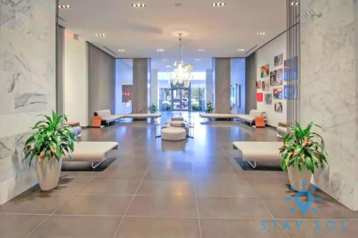 A spacious lobby with a modern design, featuring a central seating area with sofas and a chandelier. There are potted plants on either side of the seating area, and the floor is tiled.