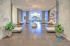 A spacious lobby with a modern design, featuring a central seating area with sofas and a chandelier. There are potted plants on either side of the seating area, and the floor is tiled. - Modern Stylish 1BR Apartment (Miami)
