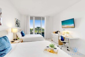 A hotel room with two beds, a desk with a chair, a large window with curtains, and a painting on the wall. - Beautiful Apt w/Private Balcony + Pool +Near Beach (Hallandale Beach)