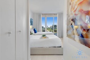 A hotel room with a large bed, a view of the outdoors, and a painting on the wall. - Beautiful Apt w/Private Balcony + Pool +Near Beach (Hallandale Beach)