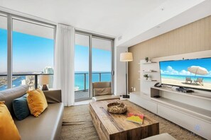 A modern living room with a large TV, a sofa, a coffee table, and a view of the ocean. - Amazing Condo + Stunning Ocean View Balcony + Gym (Hollywood)