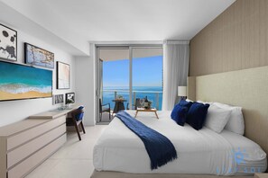 A modern hotel room with a large bed, a desk with a chair, a small table with a chair, and a balcony with a view of the ocean. - Bright & Spacious 2BR w/Balcony (Hollywood)