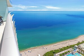 Vue panoramique d’une plage depuis un point d’observation élevé, avec une eau d’un bleu limpide, une plage de sable et un espace piscine agrémenté de parasols.