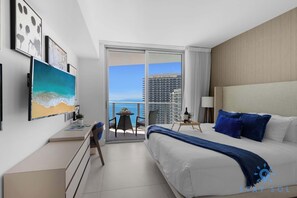 A hotel room with a bed, a desk, a chair, a small table with a bottle and glasses, and a balcony with a view of the city and ocean. - Bright & Spacious 2BR w/Balcony (Hollywood)