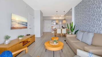 A modern living room with a sofa, a coffee table, and a TV on a wooden console. There is a kitchen in the background with a dining area.