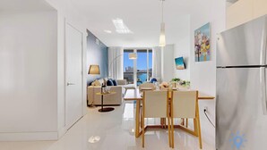 Un soggiorno moderno con divano, tavolo da pranzo e sedie, una cucina con frigorifero, e vista sulla città attraverso ampie finestre.