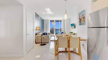 Una sala moderna con sofá, una mesa de comedor y sillas, una cocina con refrigerador, y vistas de la ciudad a través de amplias ventanas.