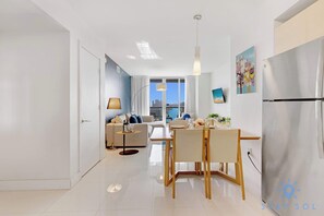 Ein modernes Wohnzimmer mit einer Couch, einem Esstisch und Stühlen, einer Küche mit Kühlschrank und Blick auf die Stadt durch große Fenster.