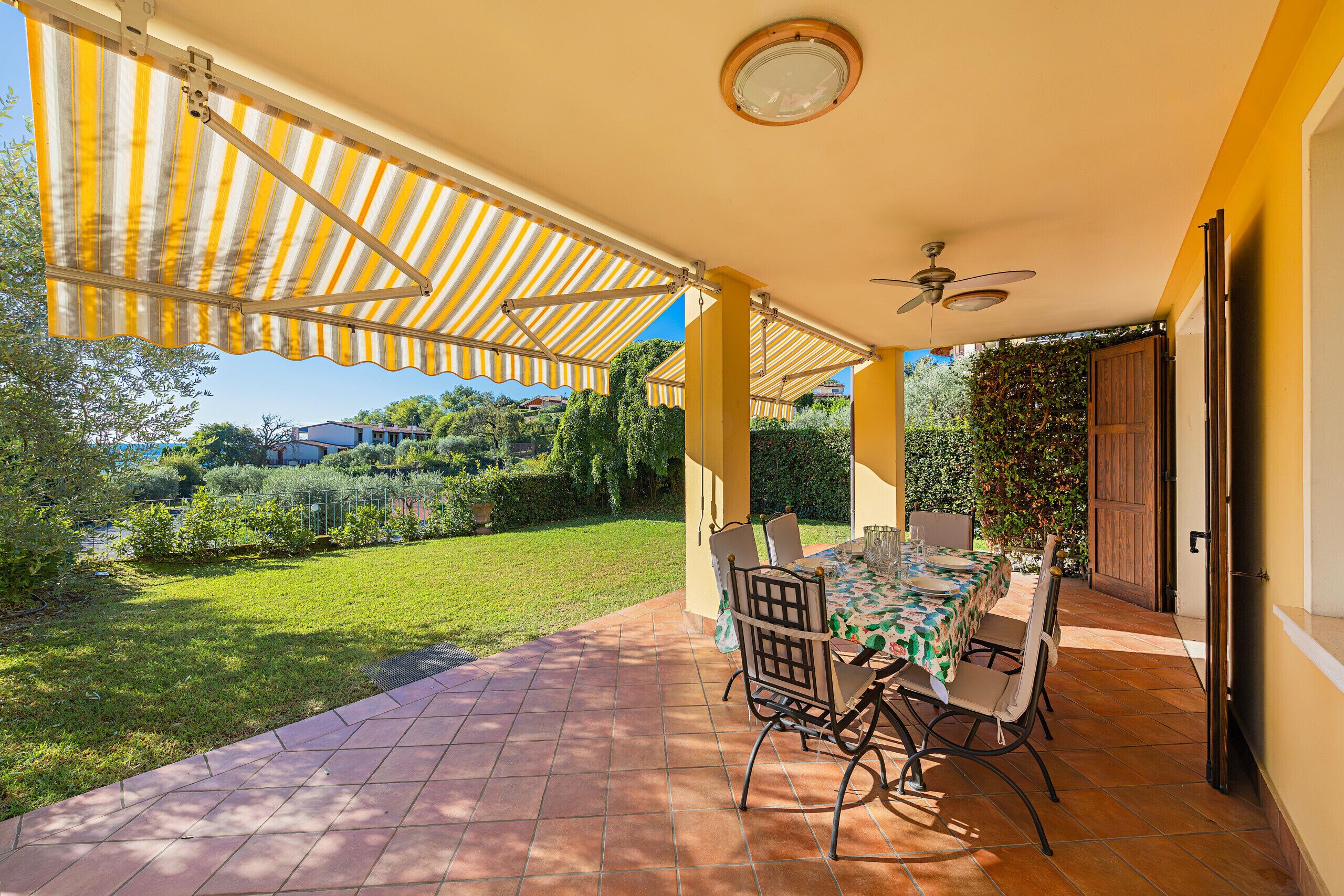 Un patio cubierto con mesa y sillas, un ventilador de techo y vistas a un jardín verde.