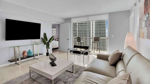 A modern living room with a flat-screen TV, a coffee table, a sofa, and a balcony with a view of high-rise buildings. - Amazing Apt w/Ocean View + Pool & Gym (Miami)