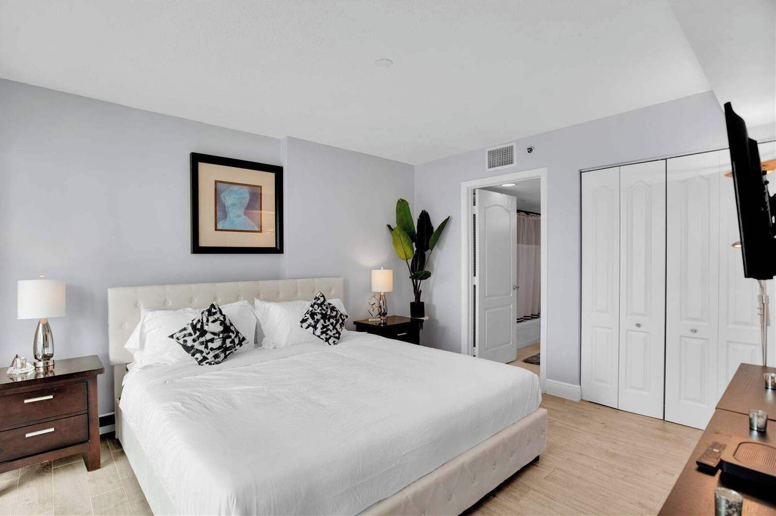 A hotel room with a large bed, bedside tables, a nightstand, a desk, a chair, a plant, and a wardrobe.