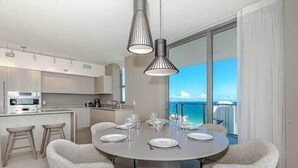 Ein moderner Essbereich mit einem runden Tisch, gedeckt für sechs Personen, umgeben von gepolsterten Stühlen. Eine Küche mit Einbauofen und einem großen Fenster mit Blick auf das Meer und die Stadt ist sichtbar.