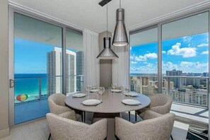 A dining area with a round table set for six, large windows offering a cityscape view, and two pendant lights above the table. - Ocean‑View 2BR Apt w/ Amazing Balcony (Hollywood)