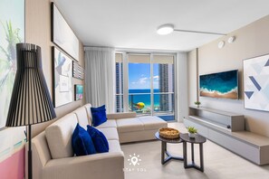 A modern living room with a sofa, a coffee table, and a TV. There is a large window with a view of a cityscape and a beach. - Beautiful 2BR Apt w/Ocean View + Pool + Amenities (Hollywood)