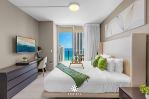 A modern hotel room with a large bed, a desk, a TV, and a balcony with a view of the ocean and buildings. - Beautiful 2BR Apt w/Ocean View + Pool + Amenities (Hollywood)