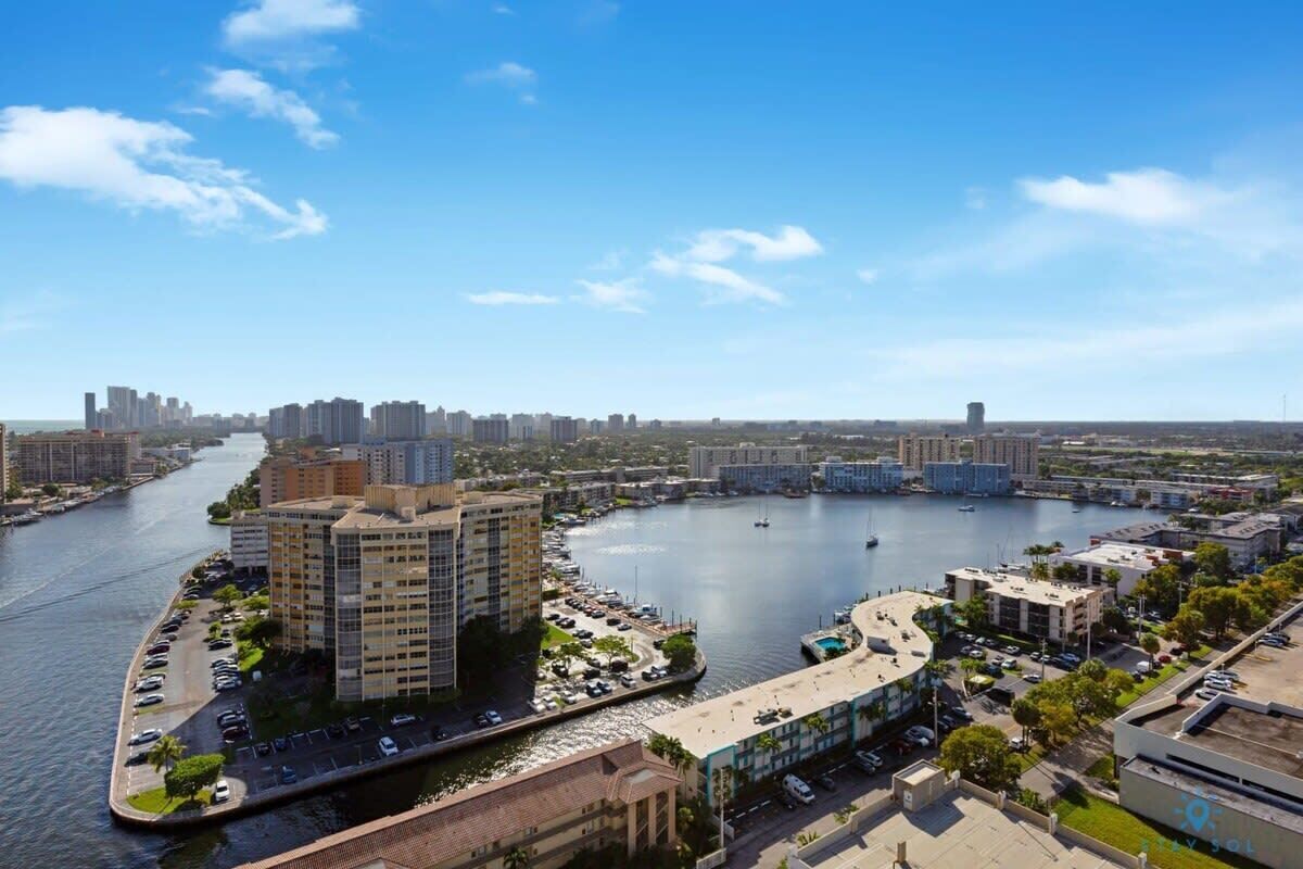 A waterfront cityscape with high-rise buildings, a marina, and a parking area.