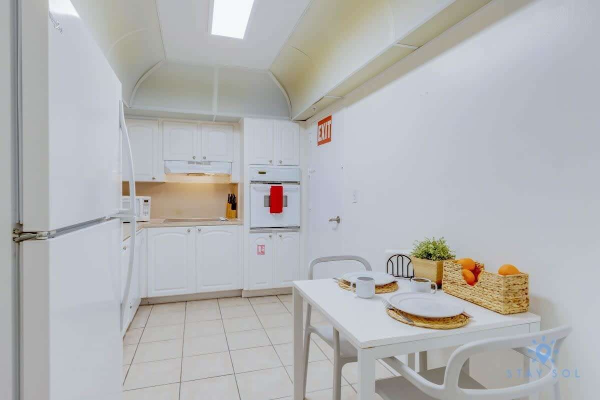 A compact kitchen with white cabinets, a microwave, and a refrigerator. There's a dining table with a basket of oranges and a small plant.