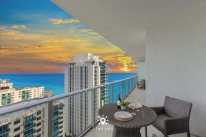 Een balkon met uitzicht op de zonsondergang boven de oceaan, een eettafel met stoelen en een fles wijn.