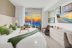 Een moderne hotelkamer met een groot bed, een balkon met uitzicht op de zonsondergang, een bureau en kunst aan de muren.