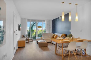 A modern living room with a sofa, coffee table, and dining table. There are three pendant lights above the table. - Miami!  Chic Apt | Pool & Balcony (Hallandale Beach)