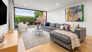 A modern living room with a grey sofa, a coffee table, and a TV on a wooden stand. There is a large window providing a view of the outdoors.